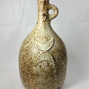 Op de afbeelding: Een bespikkelde, keramische fles met een handvat en een kurk. De fles heeft een lichtbruine basiskleur met donkerdere bruine spikkels. Een wit, reliëf ontwerp staat op de voorkant. De fles is ongeveer 18 cm hoog.