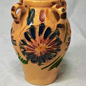 May include: A tan ceramic vase with two handles, featuring a hand-painted floral design in blue, red, and green. The surface has a textured, carved appearance with a central flower motif and brown striped accents on the handles and rim.
