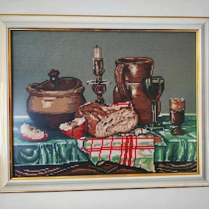 May include: A framed needlepoint still life featuring a brown ceramic pot, a loaf of bread, a sliced apple, a candle in a holder, a brown pitcher, and a glass of red wine on a green and red plaid tablecloth against a gray background.