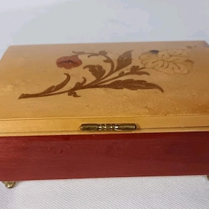 Könnte beinhalten: Eine rechteckige Schmuckschatulle aus Holz mit rotem Sockel und einem hellen, gewölbten Deckel. Der Deckel ist mit einer floralen Intarsie in Braun- und Rottönen verziert. Die Schatulle hat kleine, goldfarbene Füße und ein Messing Scharnier.