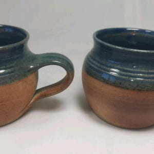 May include: Two handmade ceramic mugs. Each mug has a rounded shape with a handle. The top and handle are a dark blue-green, while the bottom is a warm brown. The mugs are suitable for coffee, tea, or other beverages.