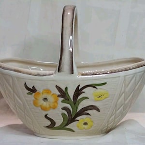 May include: Cream ceramic basket with a handle, decorated with yellow and green floral motifs. The basket has a woven pattern and a brown trim. A decorative item for the home, with a handle for carrying.