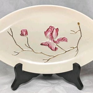 May include: An oval-shaped cream-coloured ceramic dish featuring a central hand-painted design of a pink magnolia flower and a smaller bud on thin, delicate gold-coloured branches. The dish is displayed on a black plastic stand.