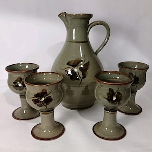 May include: A ceramic pitcher and four matching goblets. The set is a muted green with brown floral accents. The pitcher has a handle and spout. The goblets have a stem and a wide rim. The set is likely for serving beverages.