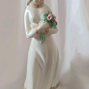 May include: A porcelain figurine of a woman in a long white dress, holding a bouquet of pink roses. The figurine has a light skin tone, with a tan collar and trim on the dress. The figure is approximately 25 cm tall.