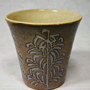 May include: A ceramic cup with a tapered shape, featuring a speckled brown exterior and a light cream-coloured interior. The outer surface is decorated with a repeating light blue botanical pattern of leaves and stems etched into the clay.