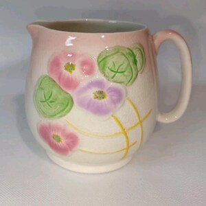 May include: A ceramic pitcher with a pink and white gradient. The pitcher is decorated with pink and purple flowers, green leaves, and yellow lines. It has a handle and a spout.