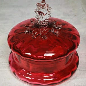 Può includere: Una ciotola per caramelle rotonda in vetro rosso intenso con coperchio abbinato. Il coperchio presenta un pomello in vetro trasparente e testurizzato al centro con un design a stella che si irradia verso l'esterno. La base e il coperchio sono lisci.