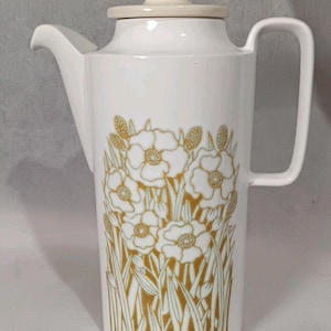 May include: White ceramic coffee pot with a lid and handle. The pot features a floral design in gold, with flowers and foliage. The lid and handle are a cream color. The pot is designed for serving coffee or tea.