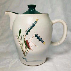 Pode incluir: Jarro de cerâmica creme com tampa e alça verde escuro. O jarro é decorado com um desenho floral em tons de verde, vermelho e amarelo. O jarro tem um bico para servir.