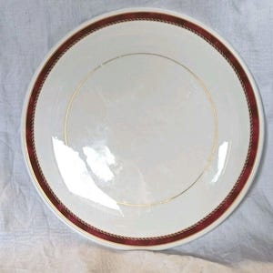 Puede incluir: Un plato de cerámica blanco con un borde burdeos y dorado. El borde presenta una banda burdeos con un diseño dorado en forma de cuerda. Una fina línea dorada rodea el centro del plato.