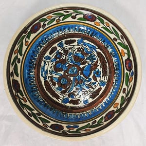 May include: A decorative ceramic bowl with a cream-colored exterior and a detailed interior design. The interior features a central floral pattern in shades of blue and brown, surrounded by a blue and brown border, and a floral border.
