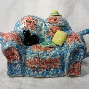 May include: A novelty ceramic teapot in the shape of a sofa, decorated with a blue, red, and white floral design. It features a black cat figurine, a yellow cushion, and a book. The lid has a hat.