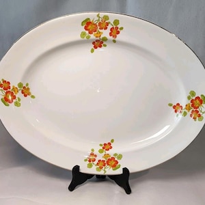 May include: Oval white serving platter with a silver rim and floral design. The platter features clusters of orange and yellow flowers with green leaves in each corner. A decorative piece for serving food.