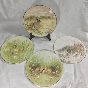 May include: Four decorative plates with gold rims. Each plate features a different wildlife scene: a rabbit in a field, two birds among flowers, two foxes, and an otter in the snow. The plates are displayed on a white surface.