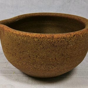 May include: A round, textured ceramic bowl with a rustic brown finish and a speckled appearance. The bowl features a distinct pouring spout on one side and a wide, open top, showcasing a deep, unglazed interior with a consistent earthy tone.