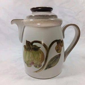 May include: Cream-colored ceramic teapot with a brown lid and handle. The teapot features a floral design in shades of green and brown. The spout is curved, and the handle is arched for easy pouring. A vintage kitchenware item.