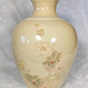 May include: A cream-coloured ceramic vase with a glossy finish and a speckled texture. The vase features a narrow neck and a rounded body decorated with a delicate, muted floral pattern in shades of soft green, brown, and pale yellow.