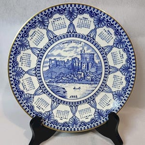 May include: A decorative plate featuring a blue and white illustration of Windsor Castle, with a calendar around the edge. The plate has a gold rim and the year 1984 is printed on the front. The plate is displayed on a black stand.