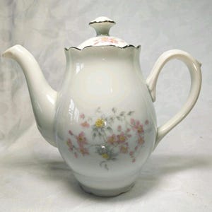 May include: White porcelain teapot with a floral design. The teapot features a curved spout, a rounded handle, and a lid with a small knob. Pink and yellow flowers with green leaves are painted on the body of the teapot.