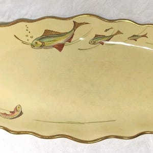 May include: An oval-shaped cream-colored ceramic serving platter with a scalloped edge trimmed in gold. The surface features a decorative illustration of several fish swimming in a line, with small bubbles rising from the largest fish.