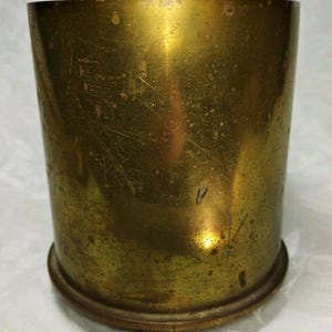 May include: A cylindrical brass container with a slightly flared base. The exterior has a tarnished, aged appearance with visible scratches and imperfections, giving it a vintage aesthetic. The container is empty.