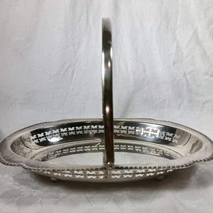 May include: A silver-colored, oval-shaped serving basket with a handle. The rim features a decorative pattern, and the basket sits on small feet. The handle is arched and attached to the sides of the basket.