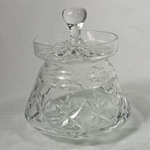 May include: A clear, cut-glass jar with a lid and a round knob. The jar has a flared base and diamond and starburst patterns. The lid is flat with a decorative edge. The knob is round and clear.