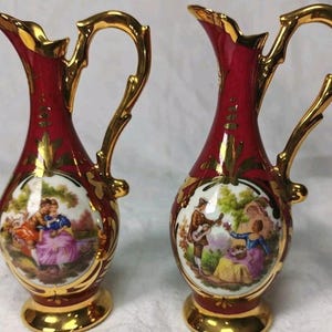 May include: A pair of matching porcelain pitchers featuring a deep red glaze with ornate gold trim on the handles, rims, and bases. Each pitcher displays a central circular medallion depicting a colourful outdoor scene with figures in period clothing.