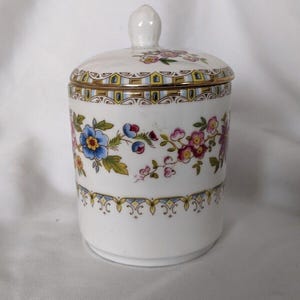 May include: A white porcelain jar with a lid, decorated with a floral pattern in shades of pink, blue, and yellow. The jar has a gold trim around the top and bottom.