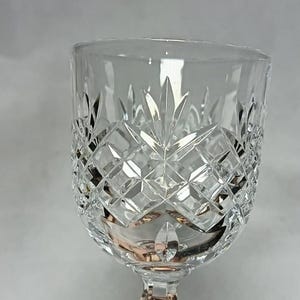 May include: A clear crystal wine glass with a diamond-cut pattern. The glass has a short stem and a wide bowl. The intricate design reflects light, creating a sparkling effect. The glass is designed for serving wine.
