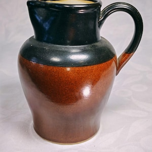 May include: A ceramic pitcher featuring a glossy black glaze on the upper portion and a rich, warm brown glaze on the lower body. The vessel has a curved handle, a wide spout, and a smooth, rounded silhouette with a distinct colour transition.