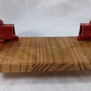 May include: A wooden workbench with two red metal vises attached. The vises are mounted on the edges of the wooden board. The wood has a natural grain pattern. The vises have silver hardware.