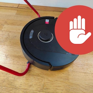 Könnte beinhalten: Ein schwarzer Roboter-Staubsauger mit einem zentralen Bedienknopf und einer roten Haltestruktur. Der Staubsauger steht auf einem Holzboden, und ein roter Kreis mit einem weißen Handsymbol befindet sich im Hintergrund.