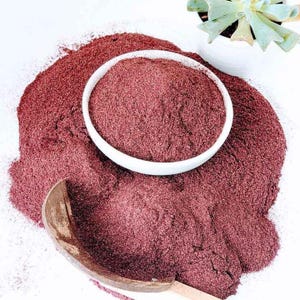 May include: A white ceramic bowl filled with fine, deep red powder, resting on a larger mound of the same dark red substance. A curved wooden scoop lies partially buried in the powder, and a small green succulent plant is visible in the corner.