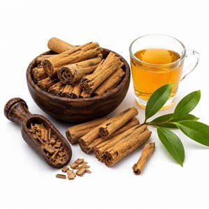 May include: A dark wooden bowl filled with brown cinnamon sticks, a matching wooden scoop containing cinnamon bark pieces, a clear glass mug filled with amber-coloured tea, and a small green leafy branch arranged on a white background.