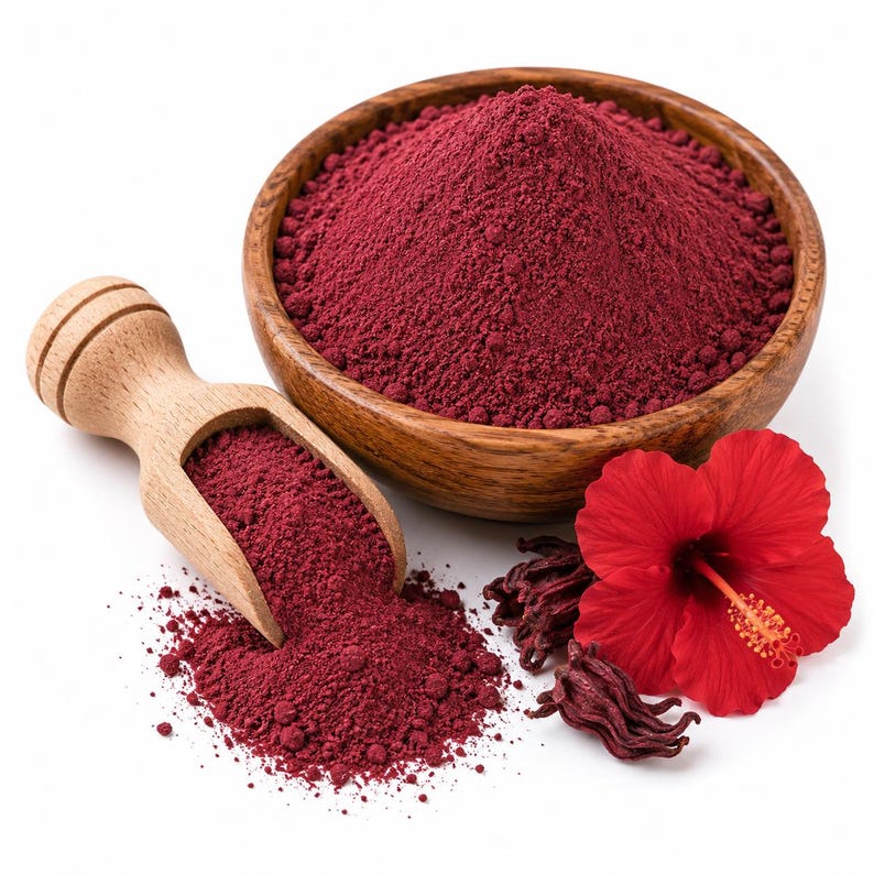 May include: A wooden bowl filled with deep red hibiscus powder, accompanied by a matching wooden scoop containing a portion of the powder. A vibrant red hibiscus flower and dried hibiscus calyces are placed next to the bowl on a white surface.
