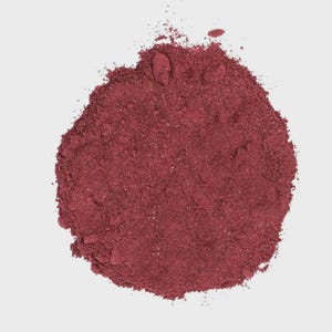 May include: A circular pile of fine, deep burgundy red powder with a dry, matte texture. The substance is granular and loosely arranged, forming an irregular mound with small scattered particles extending from the main central mass.