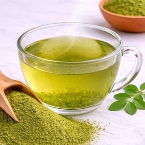 Puede incluir: Una taza de cristal transparente llena de t&eacute; verde humeante, acompa&ntilde;ada de una cuchara de madera que contiene un mont&oacute;n de polvo verde fino. Tambi&eacute;n hay un peque&ntilde;o cuenco de terracota lleno del mismo polvo verde y unas peque&ntilde;as hojas verdes.