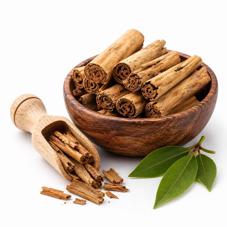 May include: A dark wooden bowl filled with light brown cinnamon sticks, accompanied by a matching wooden scoop containing smaller cinnamon pieces and fragments, alongside three fresh, smooth, pointed green leaves on a plain white background.