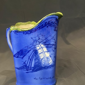 Puede incluir: Jarra de cerámica azul con una ilustración de molino de viento. El borde y la base son verdes. Las palabras "the fight with th" están escritas en la parte inferior. El asa está en el lado izquierdo. Artículo decorativo.