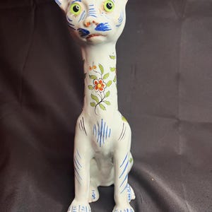 May include: A white ceramic cat figurine with blue and green floral designs. The cat has large green eyes and a long neck.