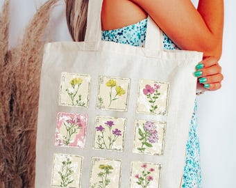 May include: A cream-colored canvas tote bag featuring a grid of nine square illustrations of various wildflowers in shades of yellow, pink, and purple. Each botanical print is framed within a distressed, weathered-looking square border.