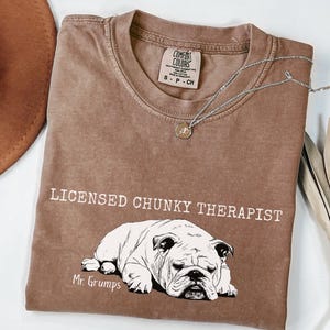 May include: A brown Comfort Colors t-shirt with the text "LICENSED CHUNKY THERAPIST" and a bulldog graphic. The shirt includes "Mr. Grumps" and a small tag at the neckline. A silver necklace with a pendant is draped across the shirt. The shirt is a small size.