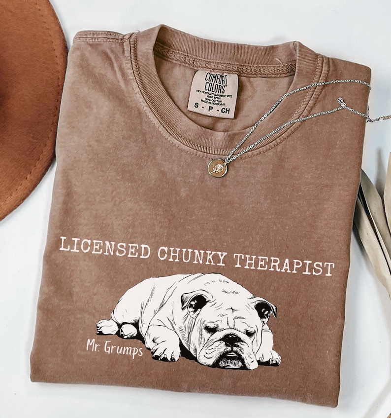 May include: A brown Comfort Colors t-shirt with the text "LICENSED CHUNKY THERAPIST" and a bulldog graphic. The shirt includes "Mr. Grumps" and a small tag at the neckline. A silver necklace with a pendant is draped across the shirt. The shirt is a small size.