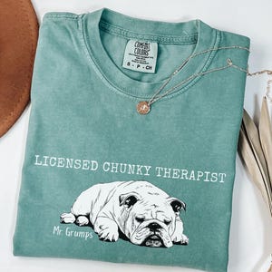 May include: A teal t-shirt with the words "LICENSED CHUNKY THERAPIST" and a black and white bulldog illustration. The shirt also features the text "Mr. Grumps" below the bulldog illustration. A gold necklace with a pendant is draped across the shirt.