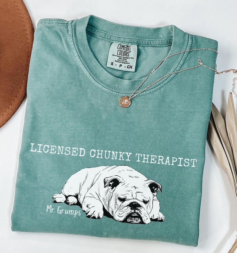 May include: A teal t-shirt with the words "LICENSED CHUNKY THERAPIST" and a black and white bulldog illustration. The shirt also features the text "Mr. Grumps" below the bulldog illustration. A gold necklace with a pendant is draped across the shirt.