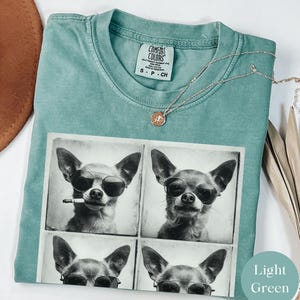 Pu&ograve; includere: T-shirt Comfort Colors verde chiaro con un collage fotografico in bianco e nero di un chihuahua con occhiali da sole, uno con una sigaretta. &Egrave; visibile anche una collana d'argento con un piccolo ciondolo.