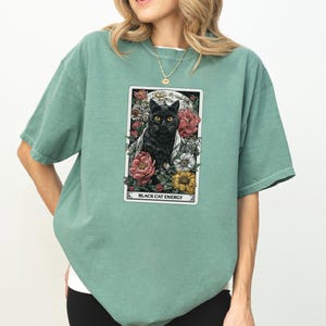 May include: Sage green t-shirt featuring a tarot card design with a black cat surrounded by flowers. The card reads "BLACK CAT ENERGY" in white text. The shirt has short sleeves and a relaxed fit.