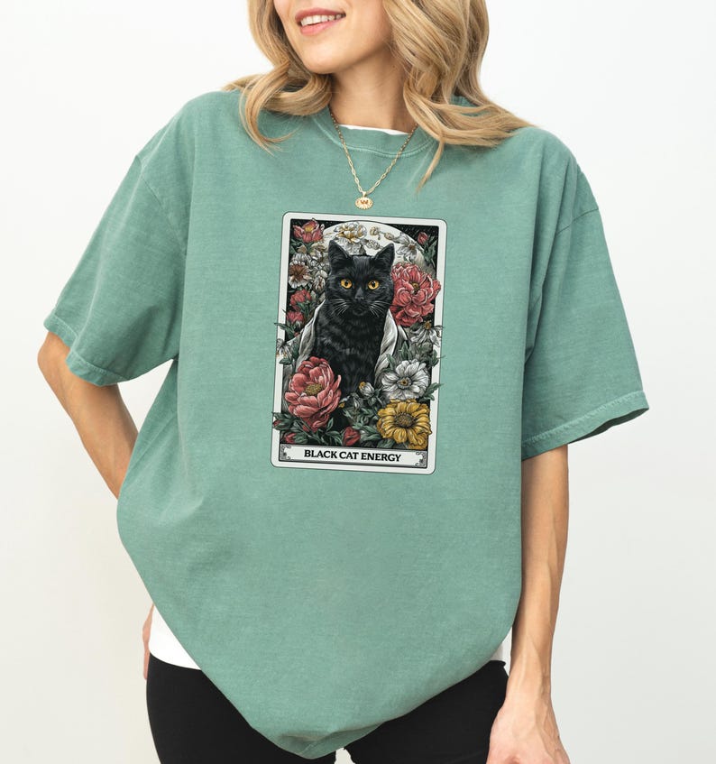 May include: Sage green t-shirt featuring a tarot card design with a black cat surrounded by flowers. The card reads "BLACK CAT ENERGY" in white text. The shirt has short sleeves and a relaxed fit.